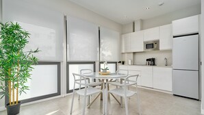 Eine moderne Küche mit weißen Schränken, einem runden Esstisch mit Stühlen, einem Topf mit Pflanze und einem großen Fenster.