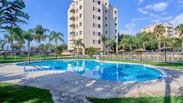 Ein Swimmingpool, umgeben von einem gepflegten Rasen, Palmen und einem mehrstöckigen Gebäude mit Balkonen.