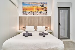 A bedroom with a bed, two nightstands, a painting of a sunset, and a door.