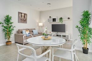 A modern living room with a round glass table, white chairs, a sofa with cushions, a flat-screen TV, and potted plants.
