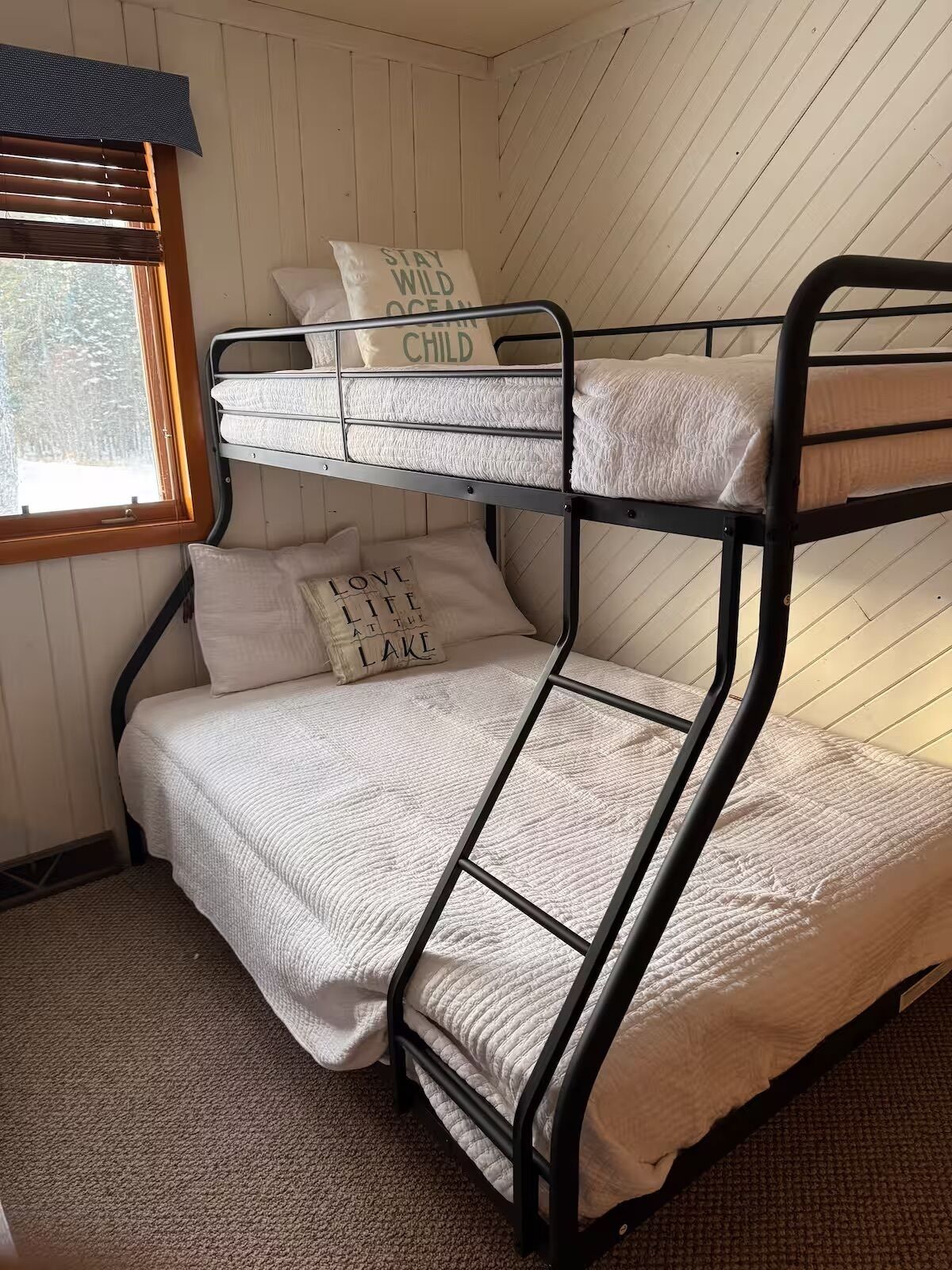 A bunk bed with a window, a pillow with "LOVE LIFE AT THE LAKE" text, and a pillow with "STAY WILD OCEAN CHILD" text.