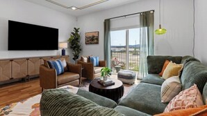 A modern living room with a large flat-screen TV, comfortable seating, a coffee table, and a view through a large window. - Family-Friendly Southside Game House (Chattanooga)