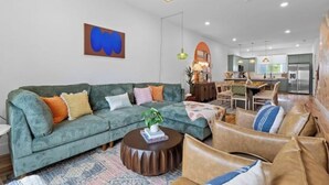 A modern living room with a green sofa, a coffee table, and a leather couch. There is a kitchen in the background with a dining area. - Family-Friendly Southside Game House (Chattanooga)