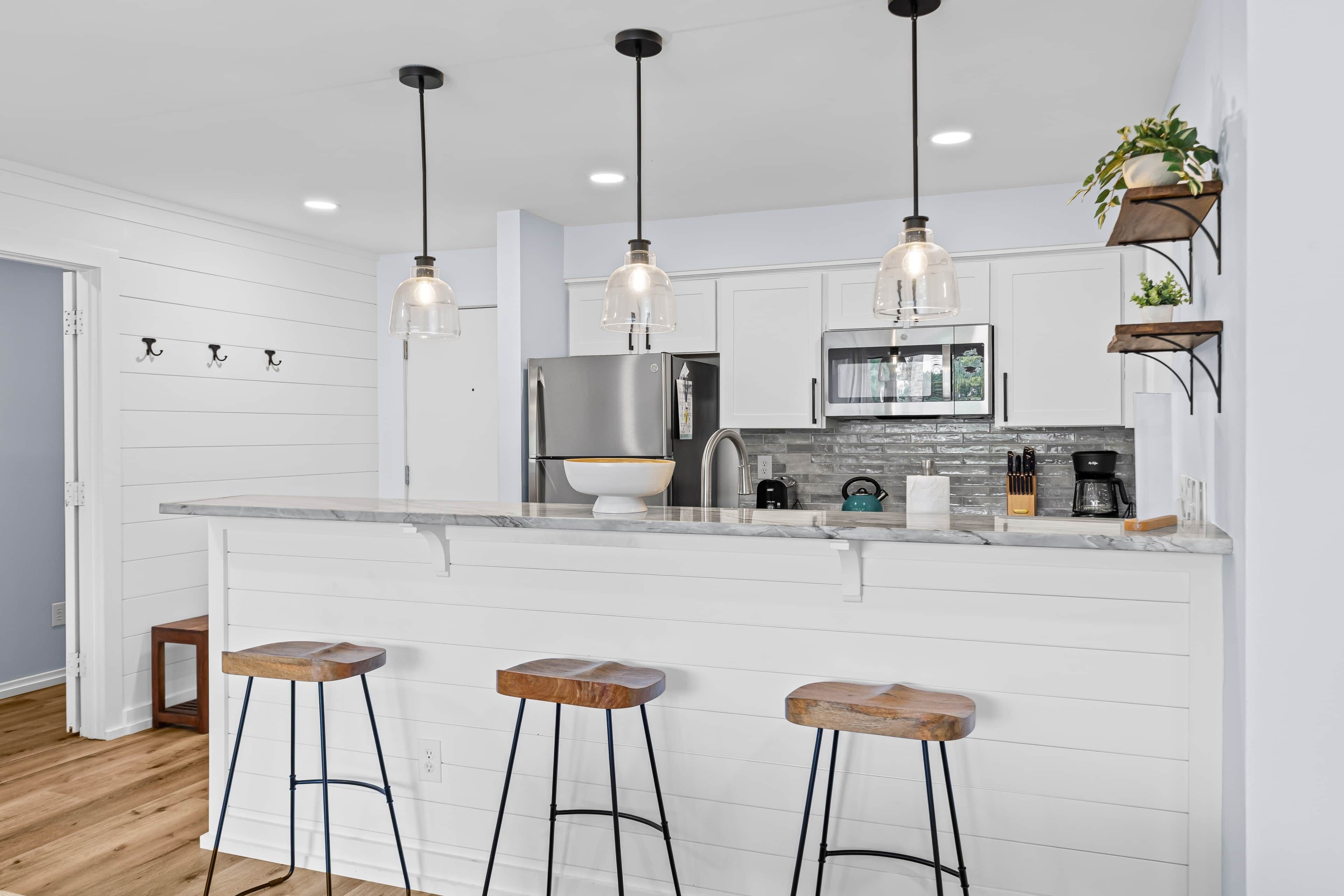 A kitchen with a marble countertop, stainless steel appliances, wooden stools, pendant lighting, and a potted plant.