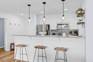 A kitchen with a marble countertop, stainless steel appliances, wooden stools, pendant lighting, and a potted plant.
