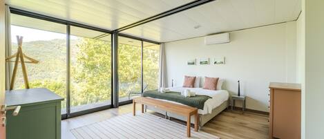 Een moderne slaapkamer met een groot raam, een bed, een bankje en een klein tafeltje.