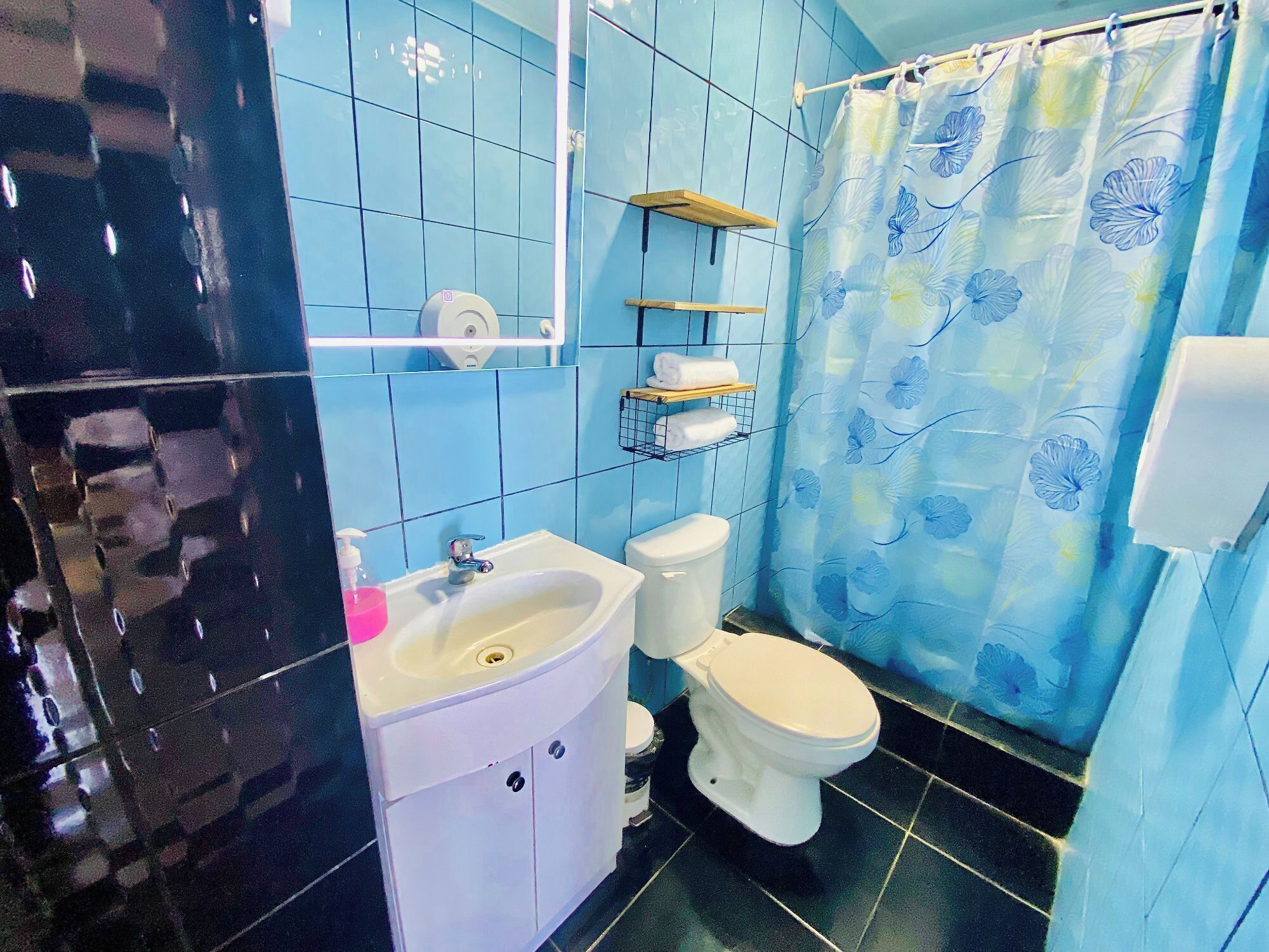 A bathroom with a white sink, toilet, and blue-tiled walls. There are shelves above the toilet and a shower curtain with a floral pattern.