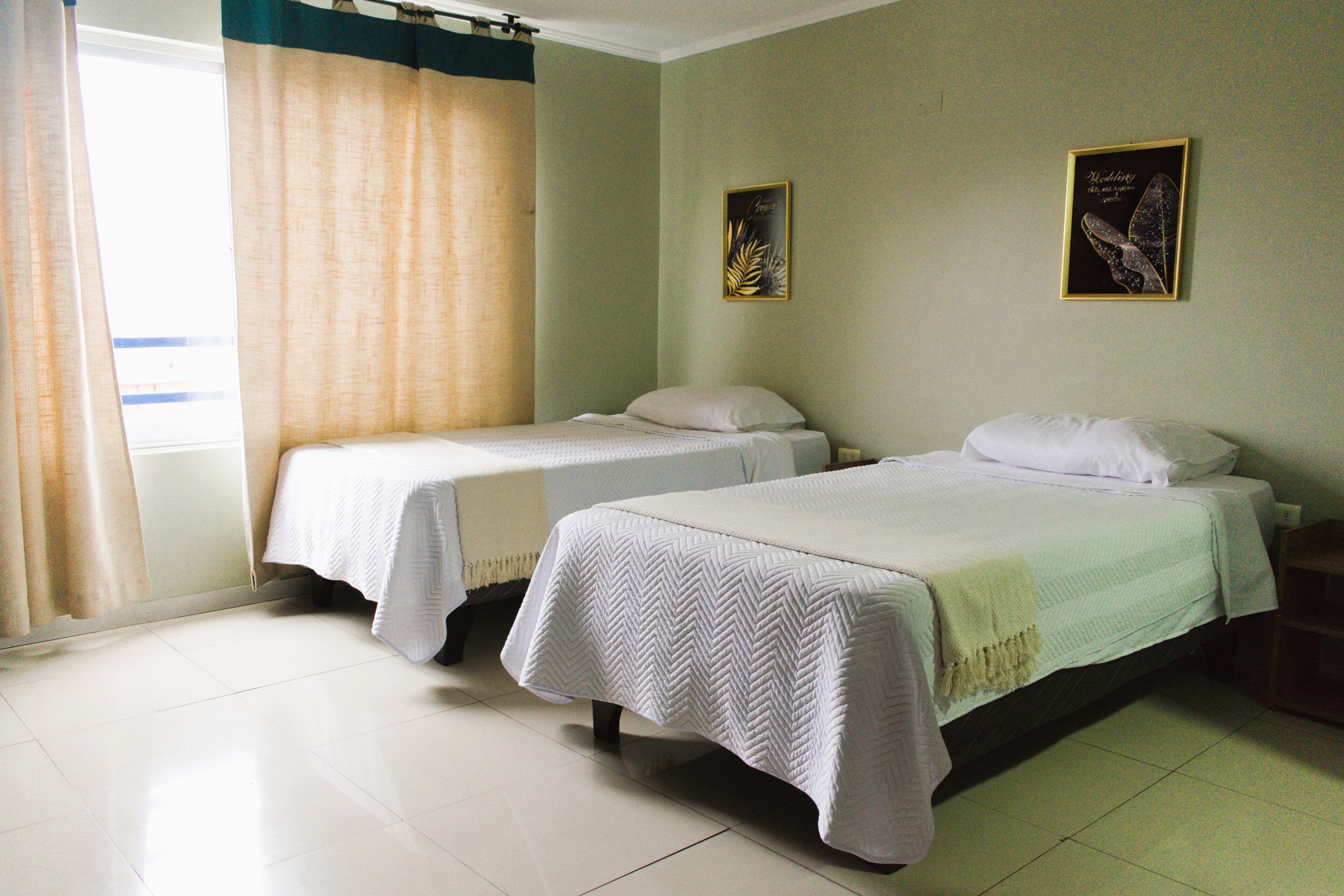 A hotel room with two single beds, a window with curtains, and two framed pictures on the wall.