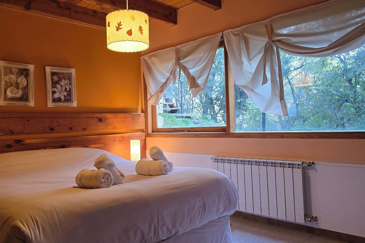 Un dormitorio con una cama grande, dos cuadros enmarcados con motivos florales, una ventana con cortinas y una lámpara colgante.