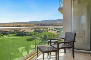 A balcony with a wicker chair, a glass railing, and a view of a golf course and buildings.