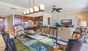A modern kitchen with a central island, dining area with a glass table and chairs, and a living room with a sofa and TV.