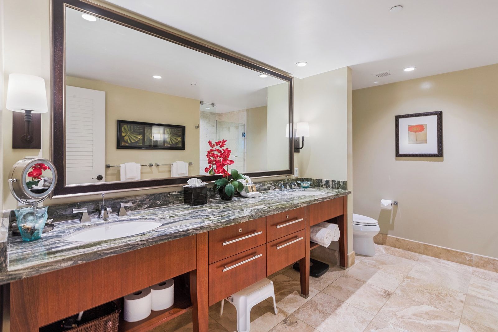 A bathroom with a large mirror, two sinks, a vanity with drawers, a trash bin, and a framed picture on the wall.