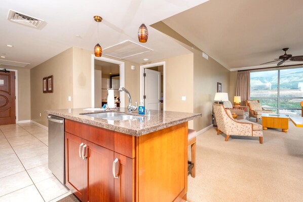 A modern kitchen with granite countertops, a built-in dishwasher, and a large island. There is a dining area with a sofa and a chair, and a large window offering a view of the city. - Honua Kai | Ocean View 7 BR, Sleeps 20 | Car Incl. w/ 6+ Nights | HKH ML-1949 by KBM (Lahaina)