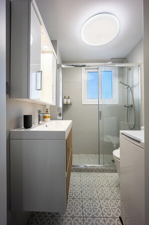 A modern bathroom with a large mirror, a white sink, a glass shower enclosure, and patterned floor tiles. - Cozy 2BR Near Metro Acropolis View (Athens)
