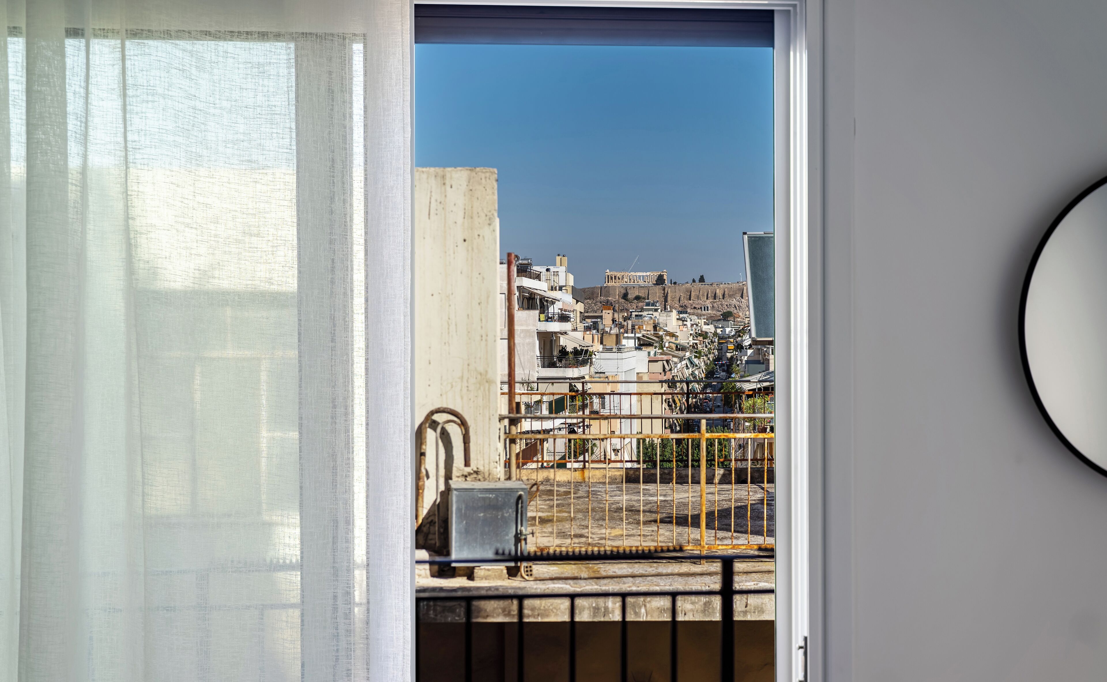 A view through a balcony door with a metal railing, a concrete pillar, and a building with a blue sky in the background.