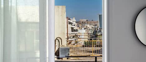 A view through a balcony door with a metal railing, a concrete pillar, and a building with a blue sky in the background.