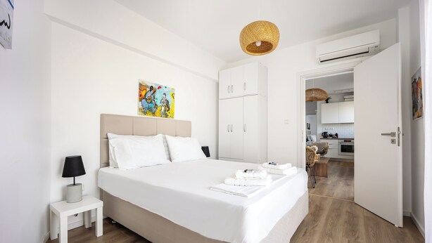 A modern hotel room with a large bed, a bedside table with a lamp, a painting on the wall, and a view into the kitchen area. - Cozy 2BR Near Metro Acropolis View (Athens)