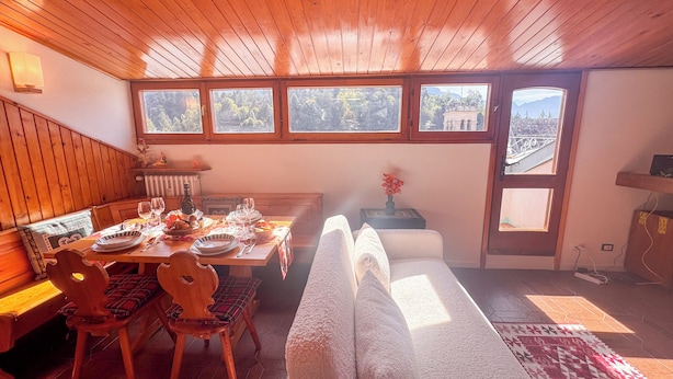 A dining area with a wooden table set for four, a bench, and a view of the outdoors through large windows. - Residenza Marmarole by Ca-Sa Check (Pieve di Cadore)