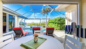 A covered outdoor seating area with a pool view, a table with a vase of flowers, and red and white chairs.