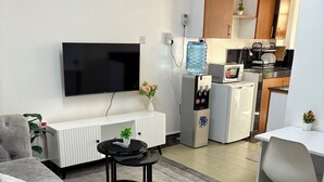 Ein modernes Wohnzimmer mit einer grauen Couch, einem schwarzen Couchtisch und einem Flachbildfernseher. In der Mitte steht ein kleiner Tisch mit einem Blumenstrauß. Im Raum befindet sich ein weißer Heizkörper, ein Wasserspender und eine Mikrowelle.
