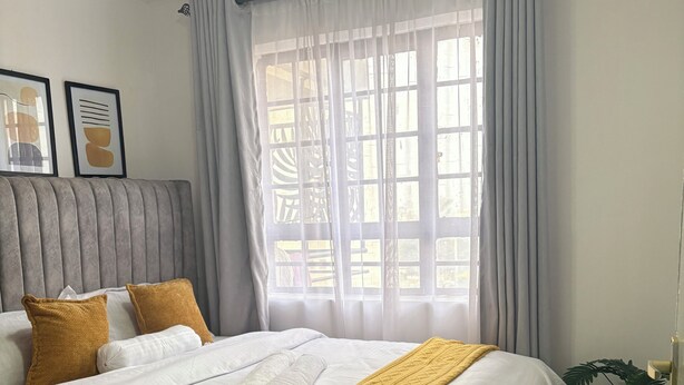 Ein ordentlich bezogenes Bett mit weißen und senffarbenen Decken, ein grauer Kopfteil und ein Fenster mit transparenten Vorhängen.