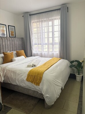 Una cama bien tendida con mantas blancas y mostaza, un cabecero gris y una ventana con cortinas traslúcidas.