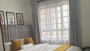 Ein ordentlich bezogenes Bett mit weißen Leinentextilien und einer gelben Tagesdecke, ein grau gepolsterter Kopfteil und ein Fenster mit transparenten Vorhängen.