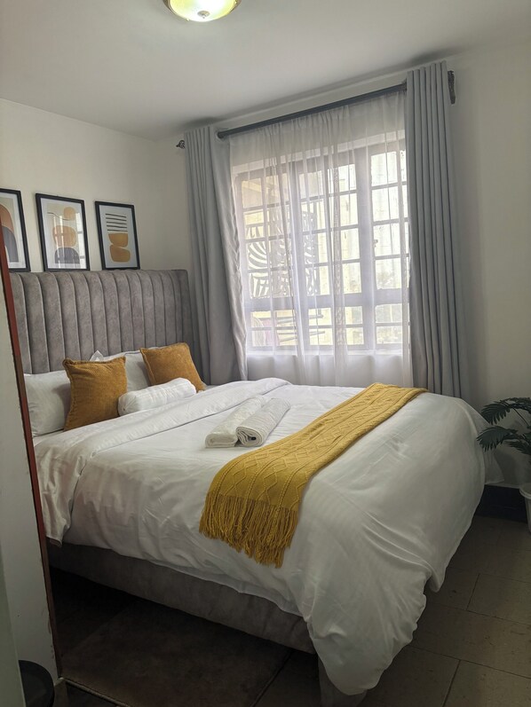 Una cama bien hecha con sábanas blancas y una manta amarilla, un cabecero tapizado en gris y una ventana con cortinas translúcidas.