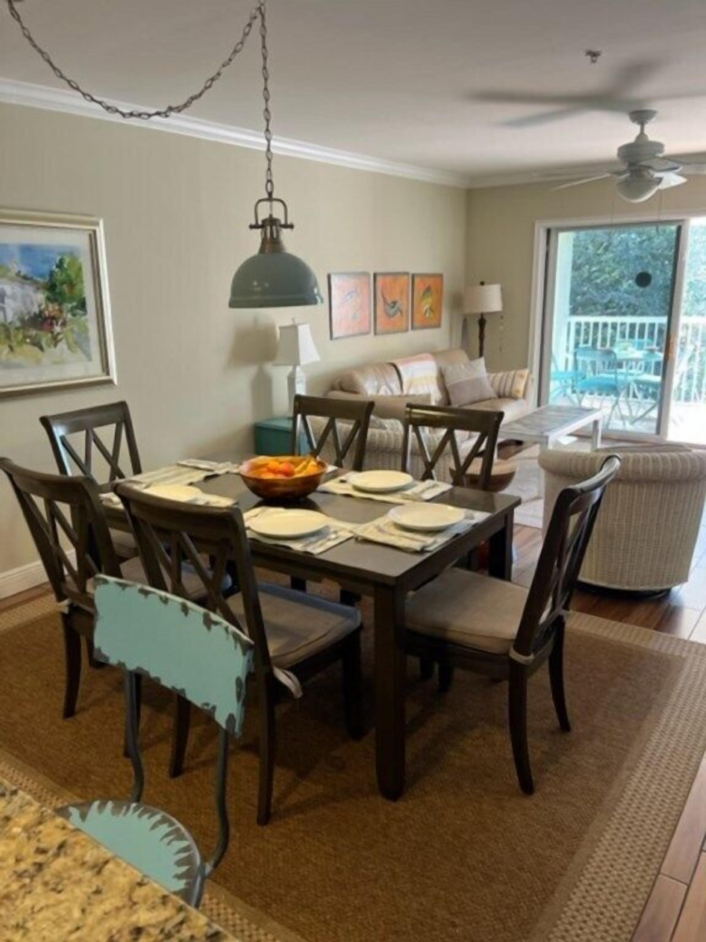 A dining area with a table set for four, surrounded by wooden chairs with cushioned seats. A hanging light fixture is above the table, and there is a large window with a view of the outdoors.