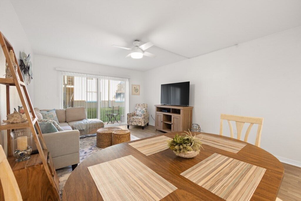 A spacious living room with a wooden dining table, a sofa, a TV, and a balcony with a view.