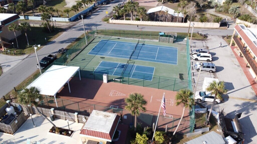 An aerial view of a tennis court surrounded by a fence, with palm trees and a parking lot in the background.