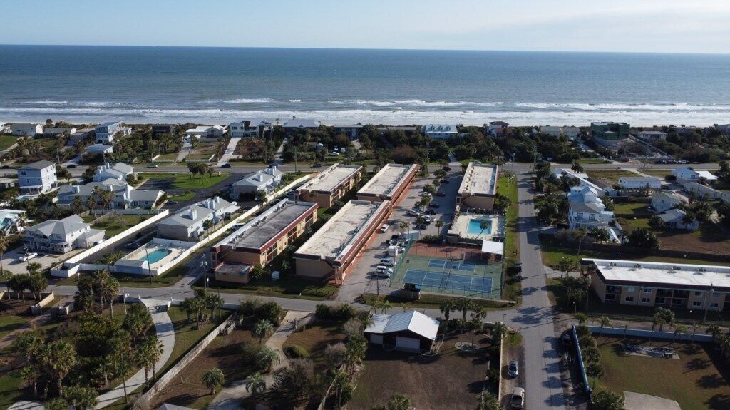Vue aérienne d’un quartier résidentiel situé sur la côte, avec des maisons, un court de tennis et une piscine.