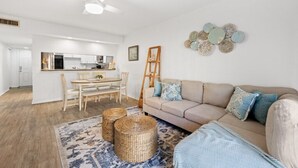 A modern living room with a beige sofa, a dining table with chairs, a kitchen in the background, and decorative wall art. - Pelican Inlet C119 - Near Beach Pool & Tennis (St. Augustine)
