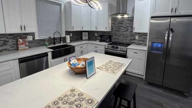 A modern kitchen with white cabinets, a stainless steel refrigerator, a built-in oven, and a large island with a bowl of snacks.
