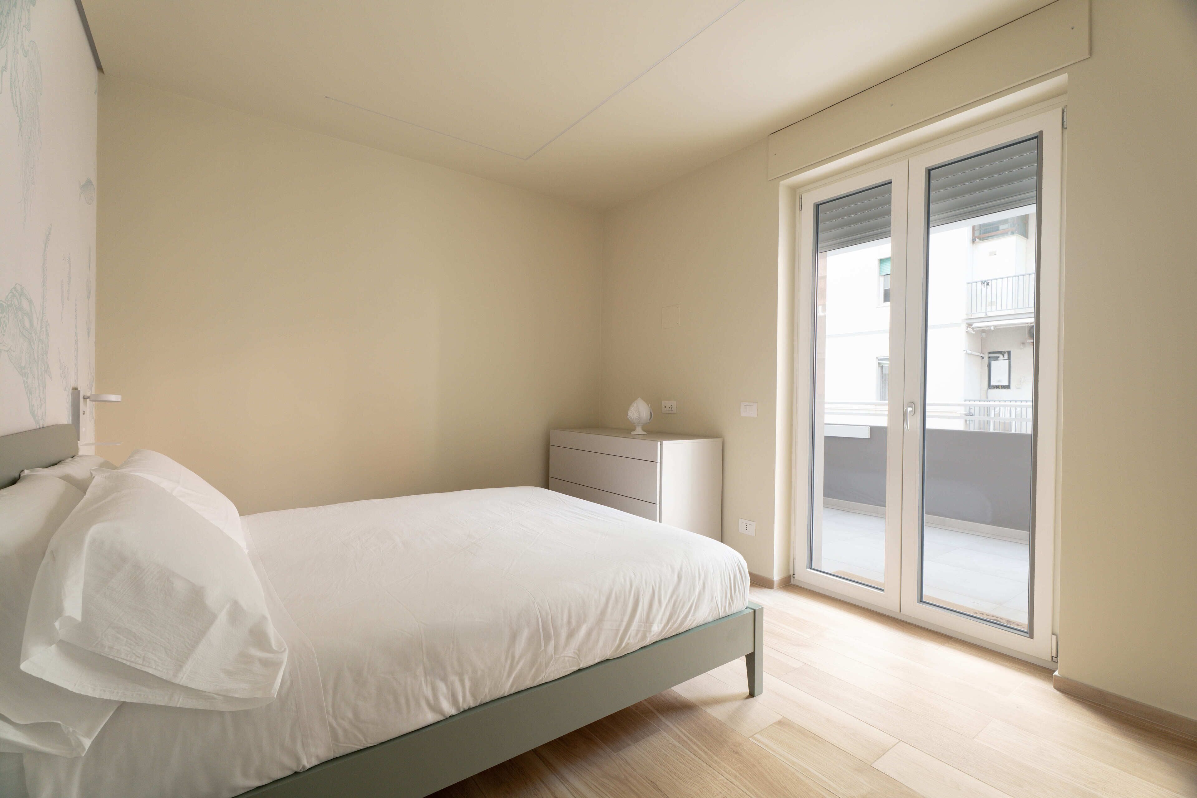 Une chambre à coucher comprenant un lit, une table de chevet, une commode et une porte-fenêtre coulissante donnant sur un balcon.