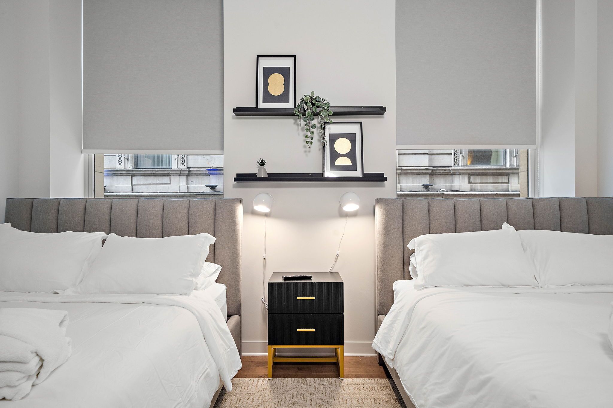 A modern bedroom with two beds, a nightstand, and a wall-mounted shelf with framed pictures and a plant.