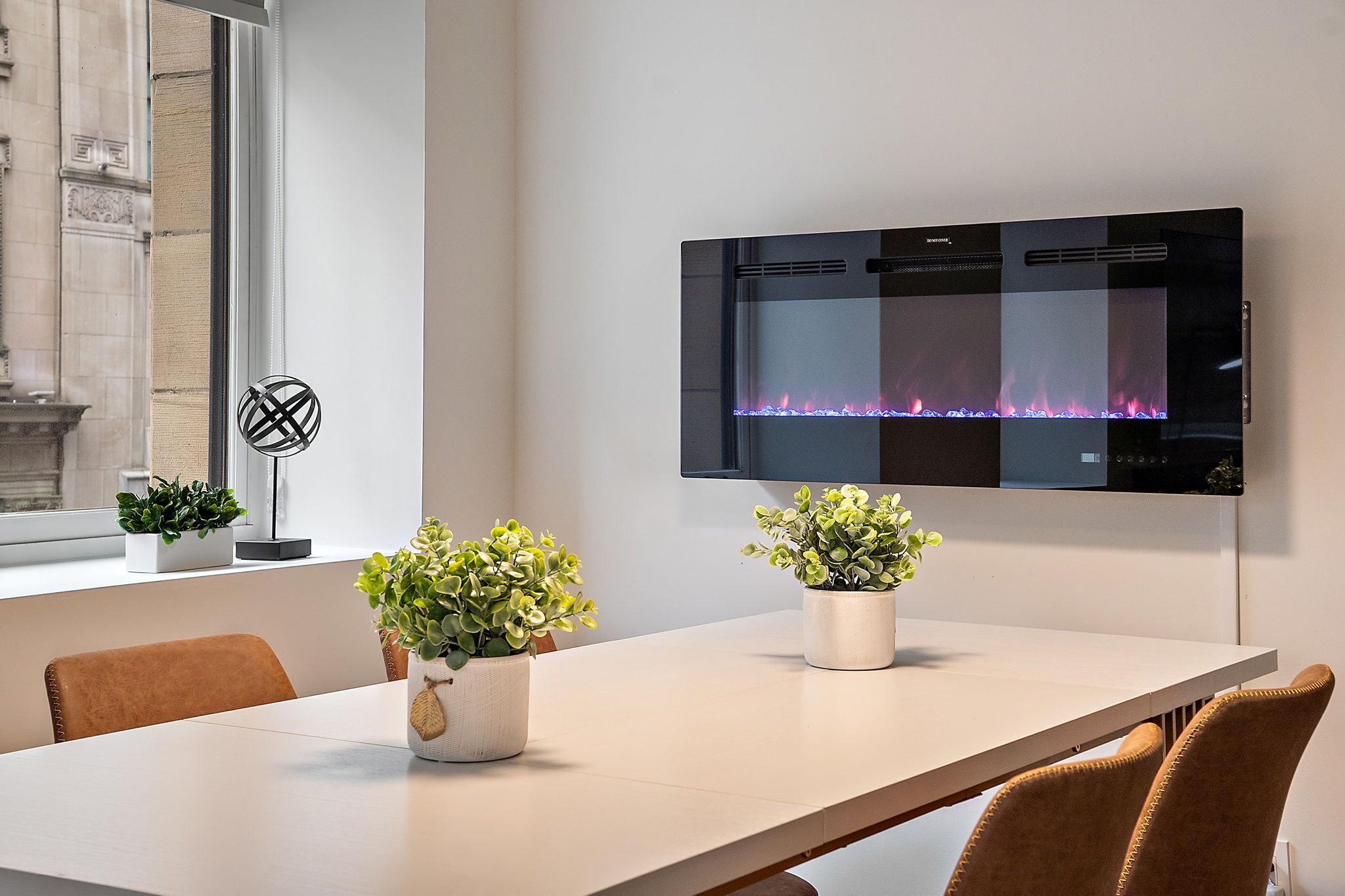 A modern dining area with a long table, two potted plants, and a wall-mounted fireplace.