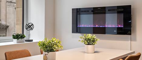 A modern dining area with a long table, two potted plants, and a wall-mounted fireplace.