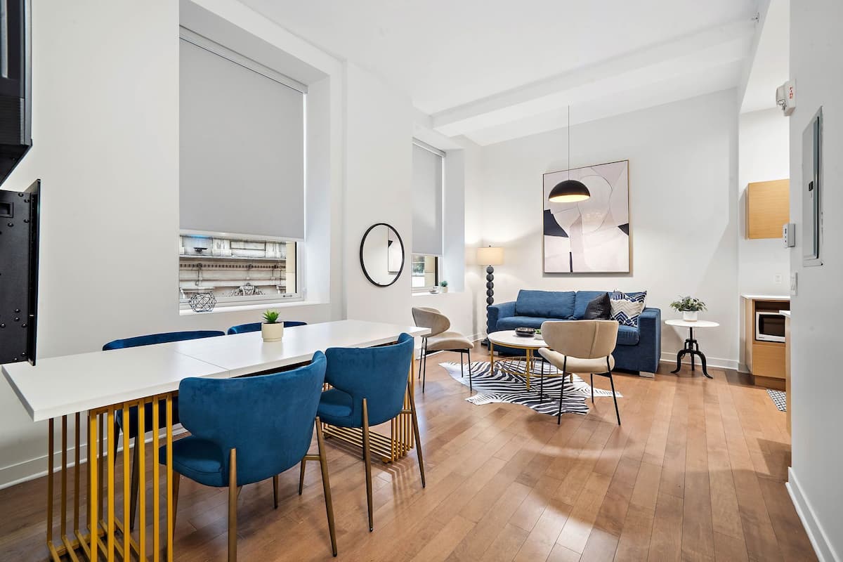 A modern living room with a dining area, featuring a white dining table, blue chairs, a blue sofa, and a small round table with a lamp.