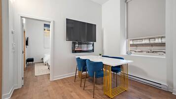 Quarto moderno com mesa branca, cadeiras azuis, televisão de tela plana e janela com vista para um edifício.