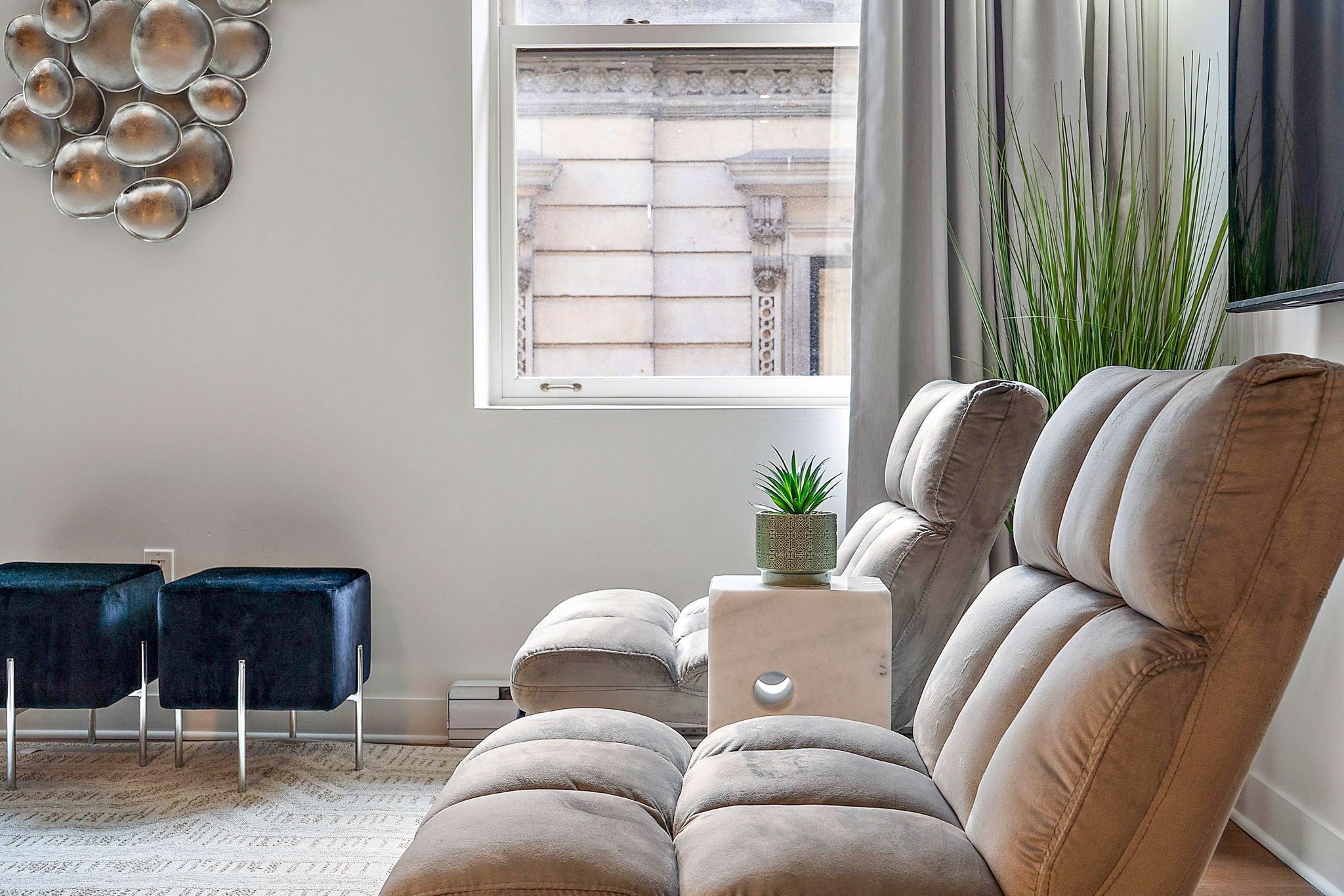 Un salon moderne avec un canapé beige, deux fauteuils bleu foncé, une petite table d’appoint avec une plante et une grande fenêtre donnant sur un immeuble.