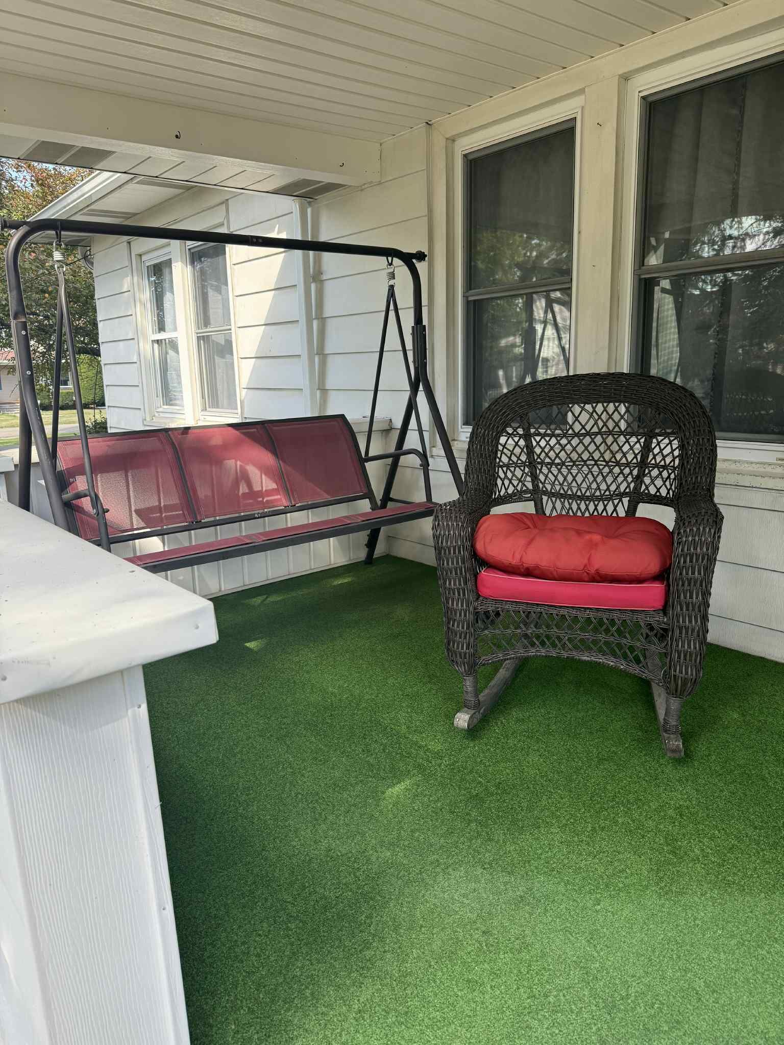 A covered porch with artificial green turf, a wicker chair with a red cushion, and a swing set.
