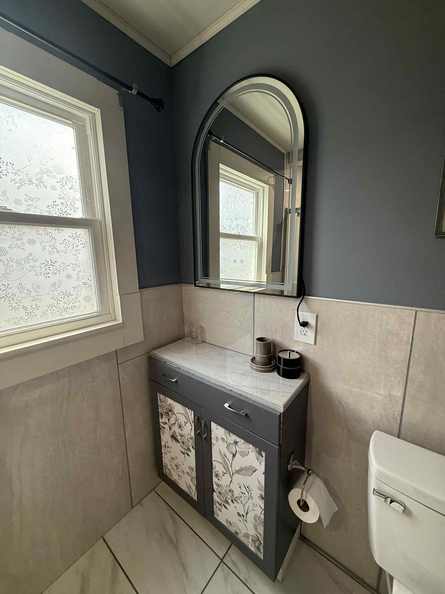 A bathroom with a dark blue wall, a white toilet, a sink with a mirror above it, and a window with floral-patterned blinds.