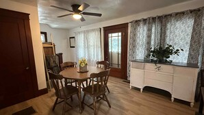 A dining area with a round wooden table, four chairs, a ceiling fan, and a window with patterned curtains.