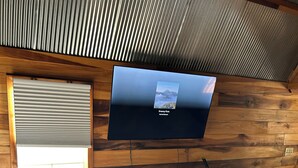 Ein Zimmer mit einer Holzwand, einem Wandfernseher, einer weißen Regaleinheit und einem Fenster mit Jalousien.