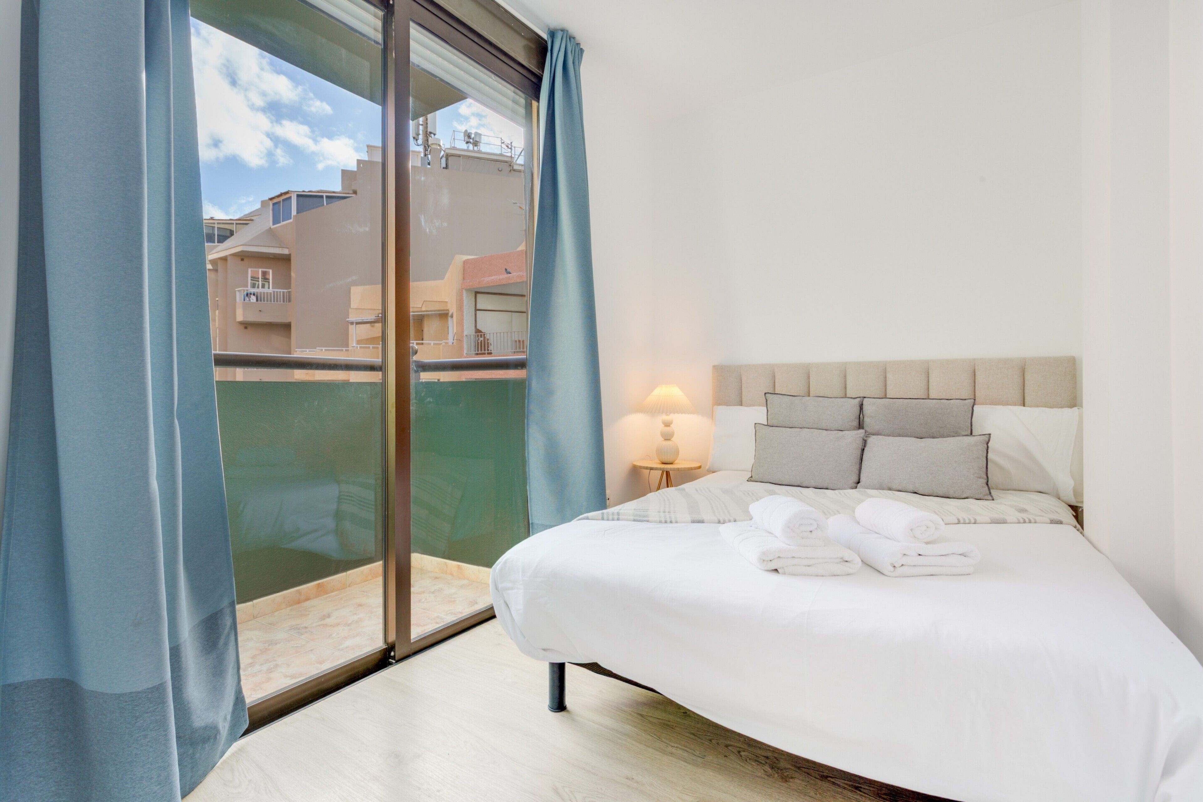 Ein Hotelzimmer mit Bett, Nachttisch, Lampe und Blick auf Gebäude durch eine Glastür.