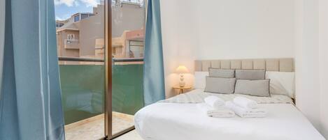 Habitación de hotel con cama, mesita de noche, lámpara y vista de edificios a través de un ventanal.