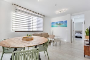 A modern living room with a round wooden table, green chairs, a beige sofa, and a painting of a beach scene.