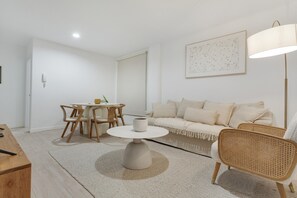 Ein modernes Wohnzimmer mit rundem Couchtisch, einer Couch, einem Korbsessel, einem Esstisch mit Stühlen und einer Stehlampe.