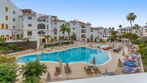 A swimming pool surrounded by lounge chairs and umbrellas, with a view of white buildings and palm trees. - Exclusive Beachfront Apartment. (Costa Adeje)